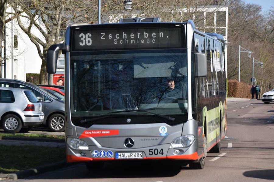 Buslinie 36 mit Zielanzeige 'Zscherben, Schmiede' fährt auf einer Straße neben parkenden Autos und Bäumen.