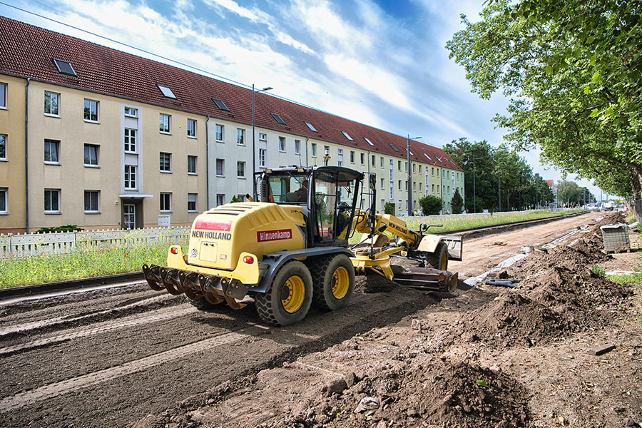 Baumaschine auf einer Baustelle neben einer langen Wohnhauszeile und Bäumen an einem sonnigen Tag.