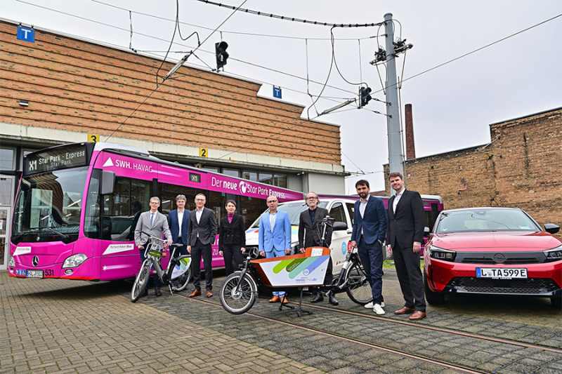 Vertreter des Projekts stehen vor den neuen Fahrrädern, Autos und einem Bus.