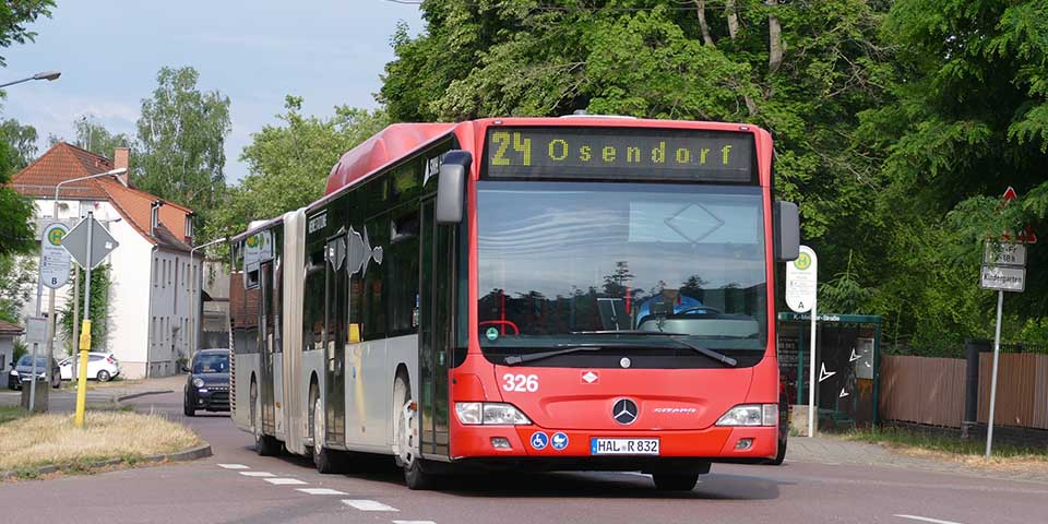 Roter Gelenkbus der Linie 24 mit Zielanzeige 'Osendorf' fährt auf einer Straße in einem Wohngebiet mit Bäumen und Häusern im Hintergrund.