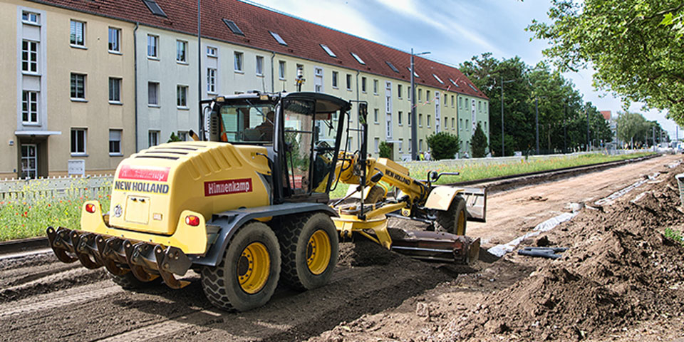 Baumaschine auf einer Baustelle neben einer langen Wohnhauszeile und Bäumen an einem sonnigen Tag.