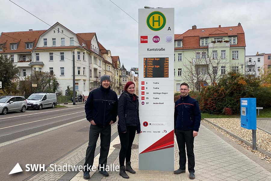 Drei Personen stehen an einer neuen Fahrgastinformationsanlage an einer Straßenbahn-Haltestelle in Halle (Saale)