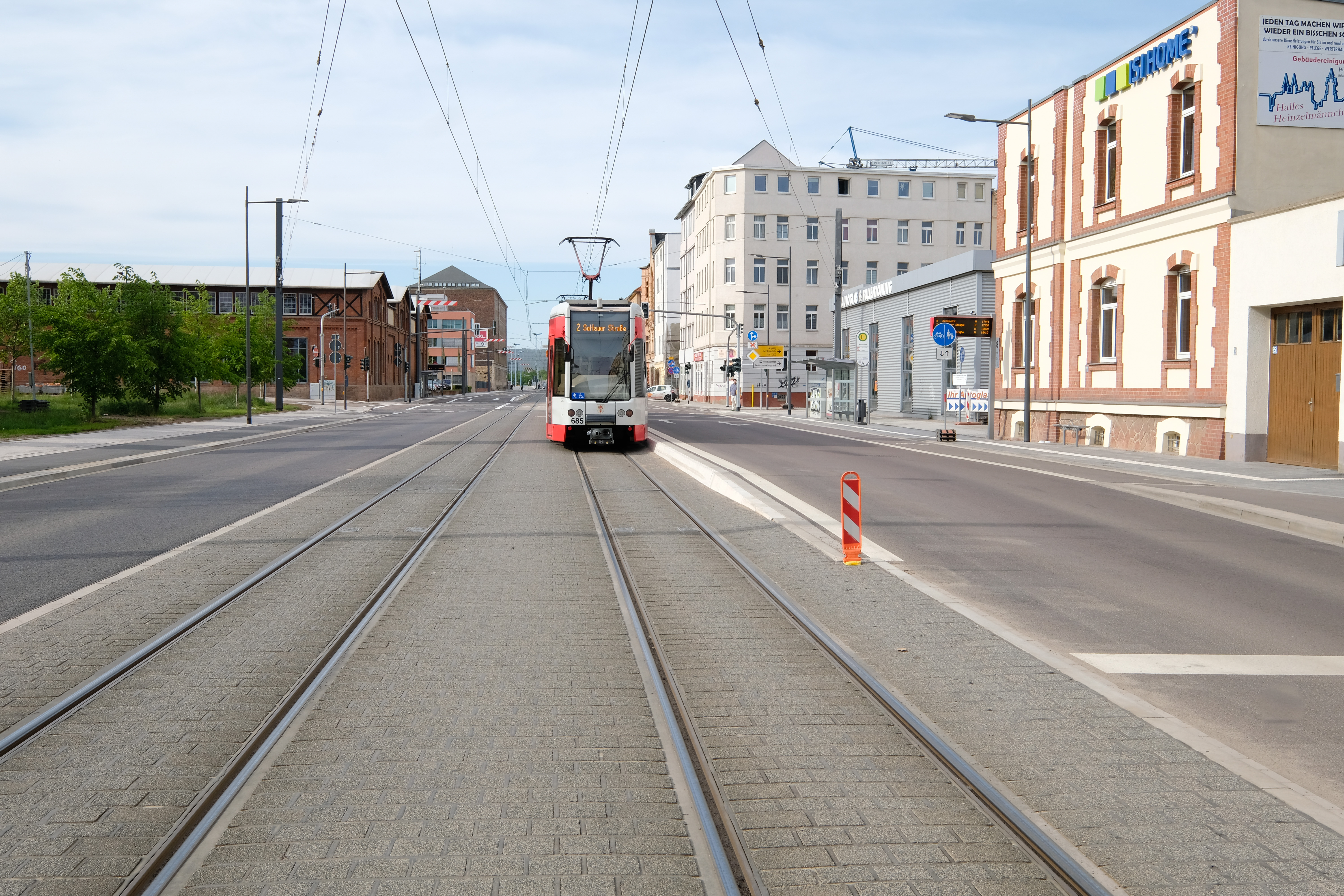 Das Bild zeigt mittig eine weiß-rote Straßenbahn auf einem Gleis, das von Pflastersteinen eingefasst ist. Sie steht an einem erhöhten Bordstein. Rechts und links der Straßenbahntrasse sind Fahrbahnen, Radwege und Fußwege.