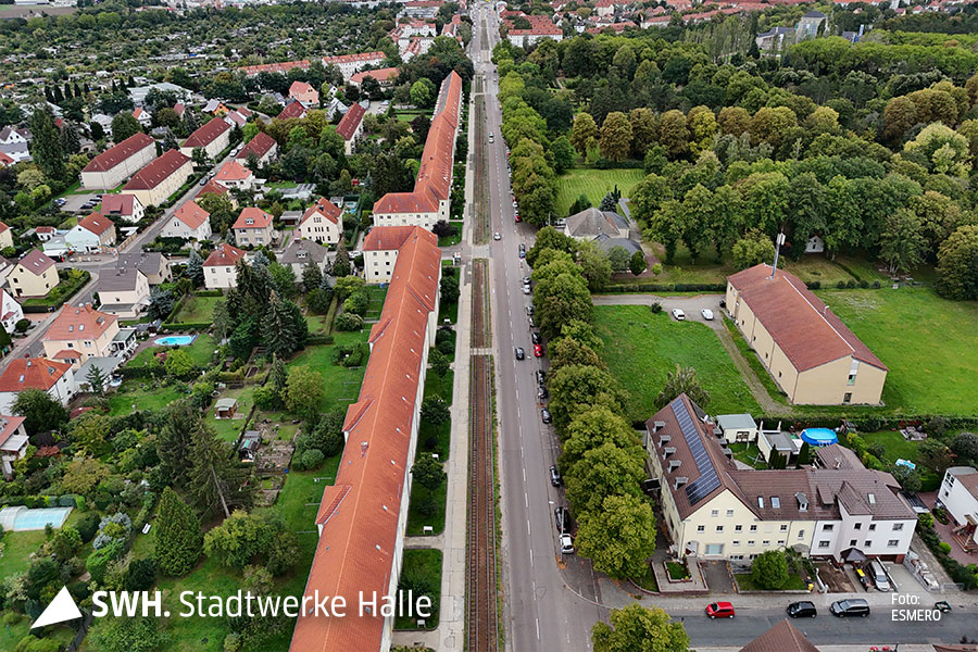 Eine Luftaufnahme der Dessauer Straße. Wenige Autos sind auf der Straße. Links neben der Straße steht ein langes Reihenhaus mit rotem Dach. Rechts stehen weniger Häuser. Mehr Grünfläche ist zu sehen.
