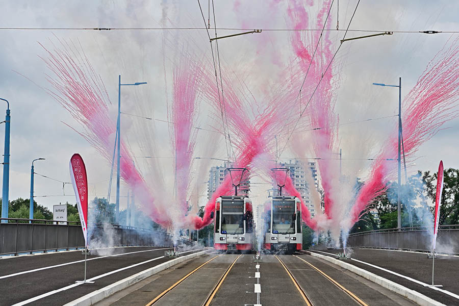 Zwei Straßenbahnen in Parallelfahrt weihen die neue Elisabethbrücke ein, im Vordergrund rot weißer Rauch und Feuerwerk.