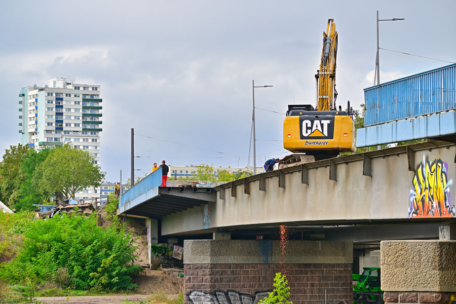 Eine Aufnahme über die saale hinweg. Ein gelber Bagger steht auf einem Stück Brücke. Das blaue Geländer fehlt an dieser Stelle.