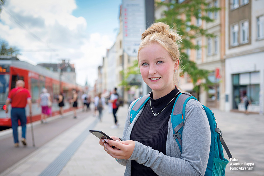 Eine Frau steht auf einem Bürgersteig in der Stadt und hält ein Smartphone in ihren Händen. Sie trägt einen Rucksack und eine graue Jacke. Im Hintergrund ist eine rote Straßenbahn und mehrere verschwommene Personen zu sehen. Die Frau lächelt in die Kamera.