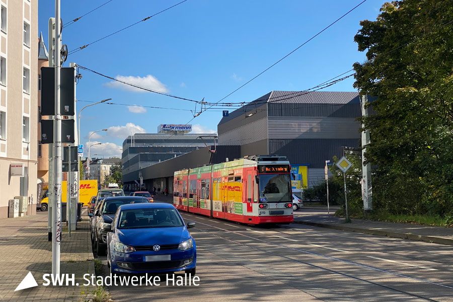 Ein Blick in die Straße: links stehen parkende Autos, ganz vorn im Bild ein blauer VW. Rehts steht eine rote Straßenbahn. Der Himmel ist blau, die Sonne scheint. Im Hintergrund sieht man ein Gebäude auf dem "Lührmann" steht.