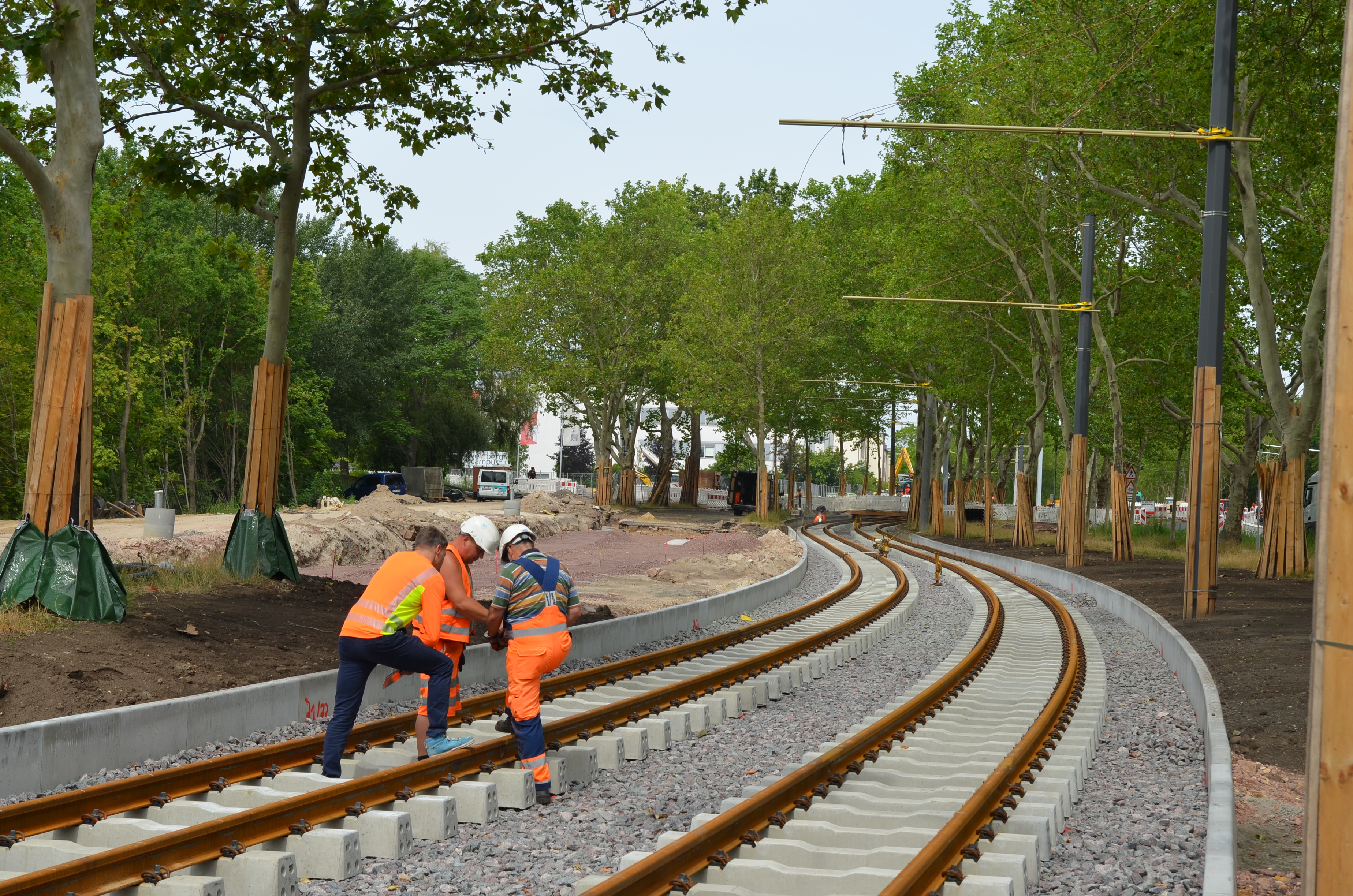 Hier sind zwei neu verlegte Straßenbahngleise zu sehen, die in einer Kurve verlegt wurden. Auf der linken Seite stehen drei Männer in orangefarbener Kleidung, die sich zu beraten scheinen. Rechts und links der Schienen stehen viele grüne Bäume.