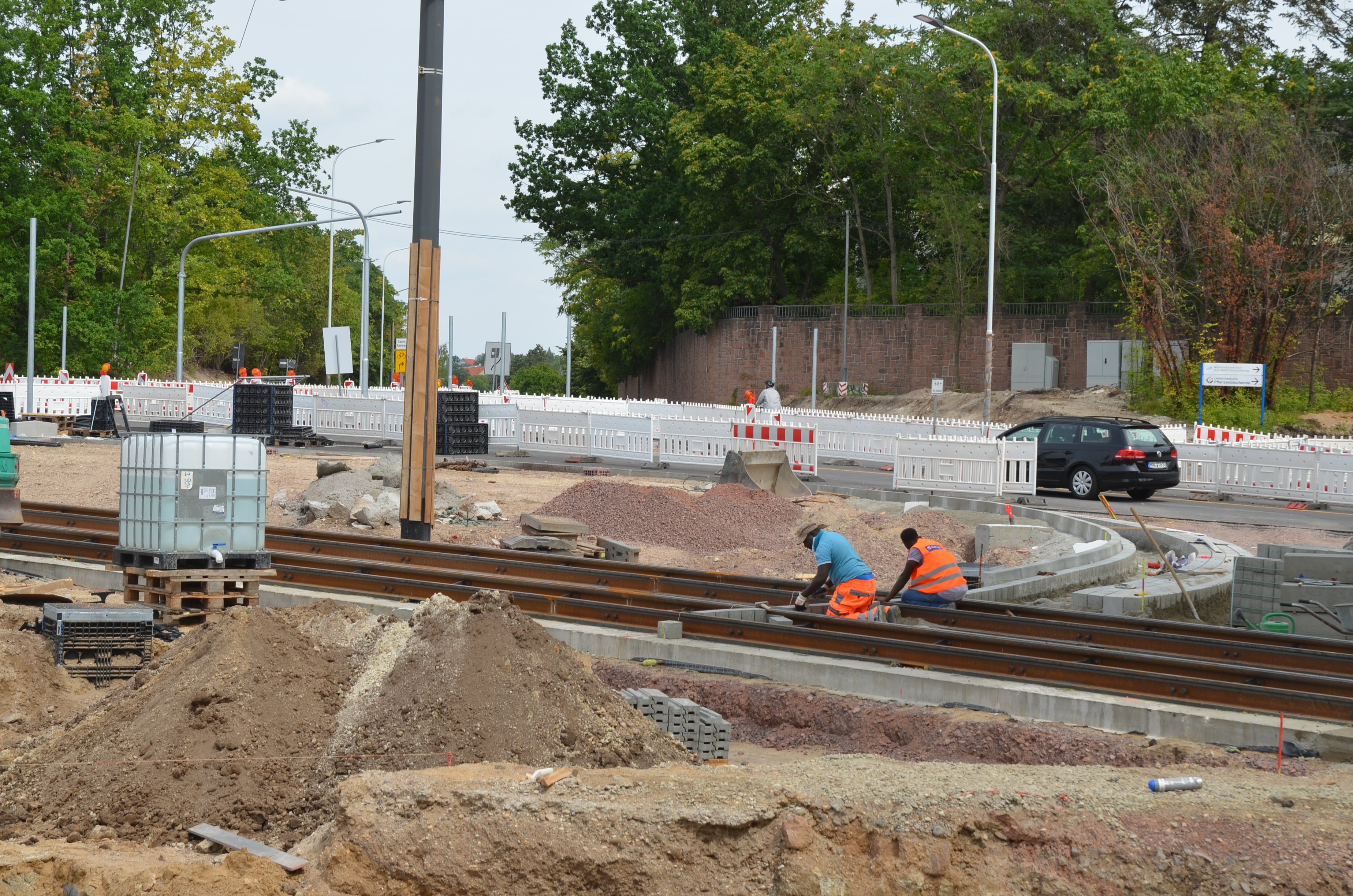 Hier ist die Baustelle am Kreisverkehr abgebildet. Von links nach rechts verlaufen Straßenbahnschienen durch das Bild, an denen zwei Bauarbeiter in orangefarbener Kleidung arbeiten. Um sie herum befinden sich aufgrund von Tiefbauarbeiten viele Sand- und Steinhügel.