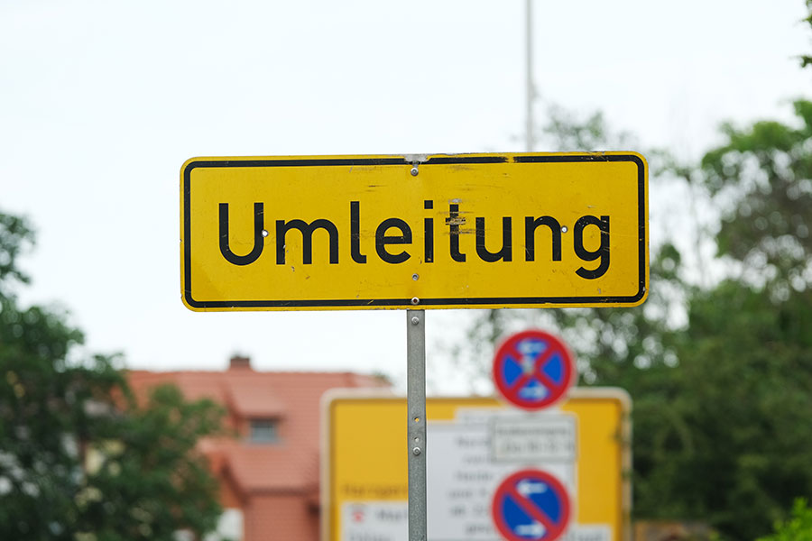 Eine Nahaufnahme eines Straßenschildes. Auf dem gelmen Schild steht mit schwarzer Schrift "Umleitung". Im Hintergrund ist ein rotes Dach eines Hauses, Bäume und trüber Himmel zu sehen.