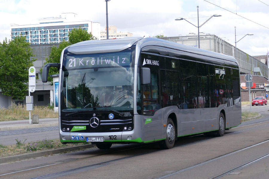 Ein silberner E-Bus der HAVAG der Marke Mercedes fährt über eine Straße, in der auch Gleise liegen. Im Hintergrund stehen vershiedene Häuser, wenige Bäume. Der Himmel ist wolkig.