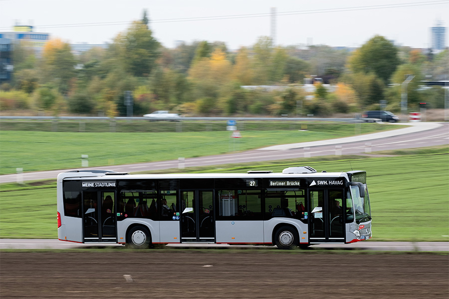 Ein silberner Bus der HAVAG fährt auf der Straße von links nach rechts durch das Bild. Im Vordergrund ist Acker zu sehen, im Hintergrund Wiese, Bäume und eine Straße. Der Fokus liegt auf dem Bus, der Hintergrund ist unscharf. 