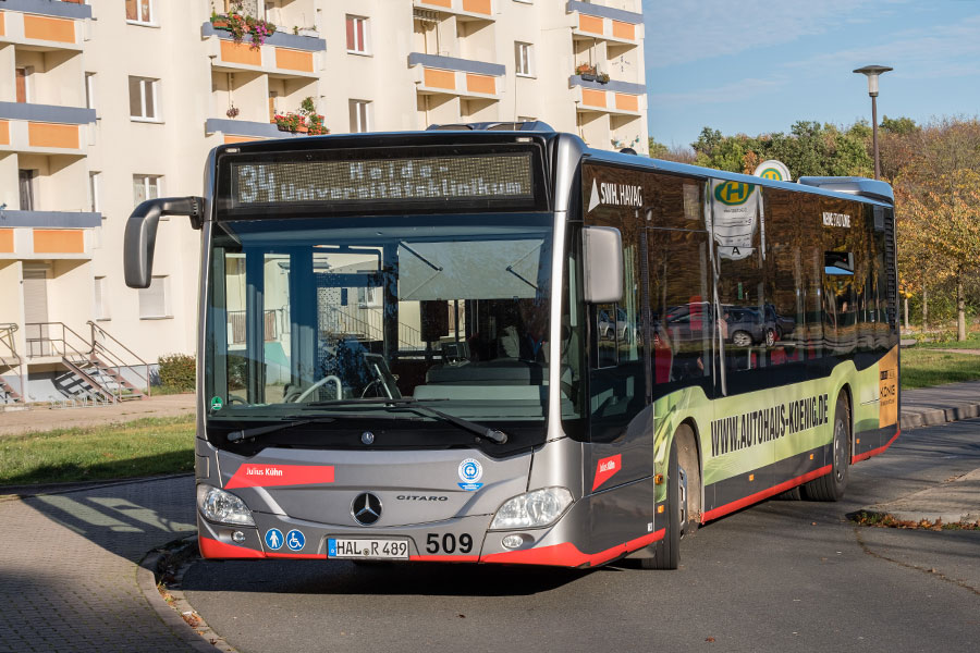 Ein silbernen Bus der HAVAG auf der Straße. Im Hintergrund ist ein Plattenbau zu sehen. Der Himmel ist blau, es ist sonnig.