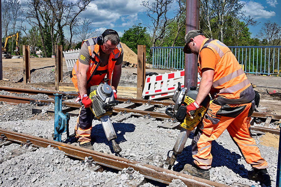 Zwei Männer in orangefarbener Arbeitskleidung stehen sich gegenüber. Sie halten jeweils einen Bohrhammer in der Hand. Sie stehen an den Gleisen.