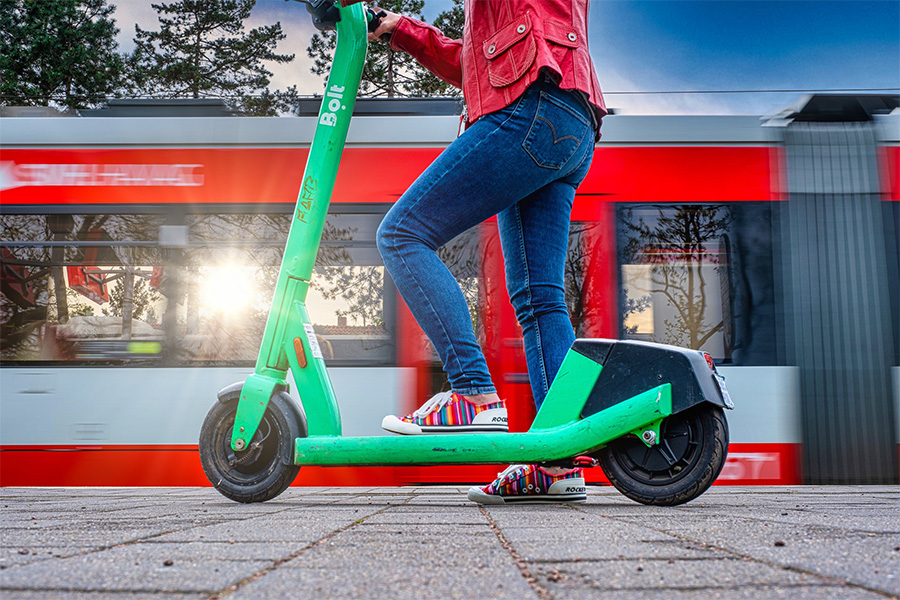 Das Bild zeigt eine Person auf einem grünen E-Scooter. Die Person trägt eine rote Jacke, blaue Jeans und bunte Sneaker. Der E-Scooter steht auf einem gepflasterten Boden, im Hintergrund fährt eine rote Straßenbahn der Stadtwerke Halle vorbei. 