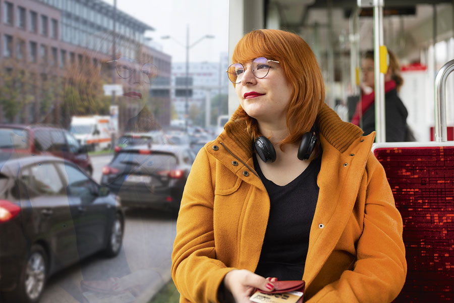 Eine Frau mit rostroten Harre, Brille und orangem Mantel sitzt in einer Straßenbahn und schaut verträumt nach draußen durch die Fensterscheibe der Straßenbahn.