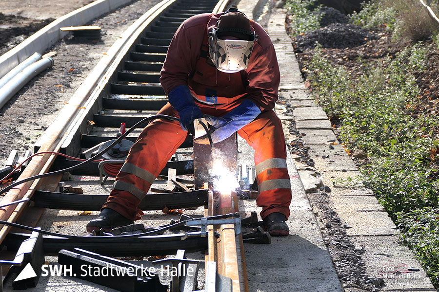 Ein Mann in orangefarbener Arbeitsbekleidung steht gebückt über einem Gleis. Er schweißt und trägt einen Gesichts- und Kopfschutz.