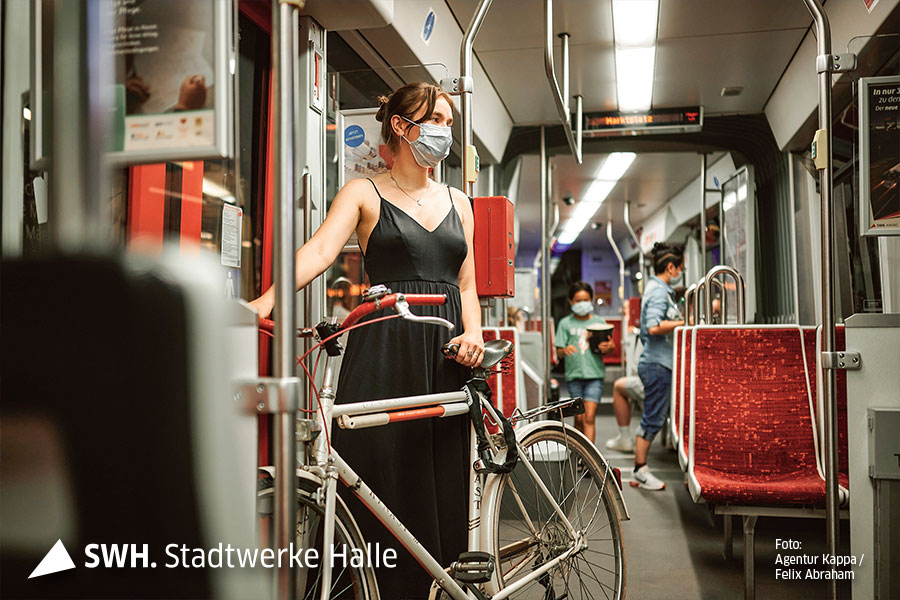 Eine Frau mit Maske und schwarzem Kleid steht mit einem sportlichen Fahrrad in der Straßenbahn.