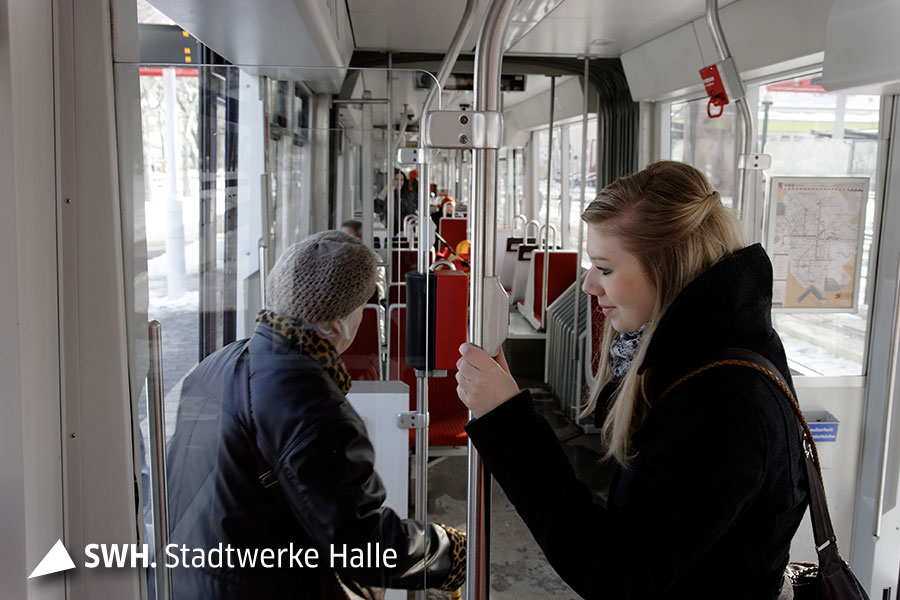 Eine junge blonde Frau hält sich an der Sicherheitsstange in der Straßenbahn fest und eine ältere Frau kommt zur Tür herein.
