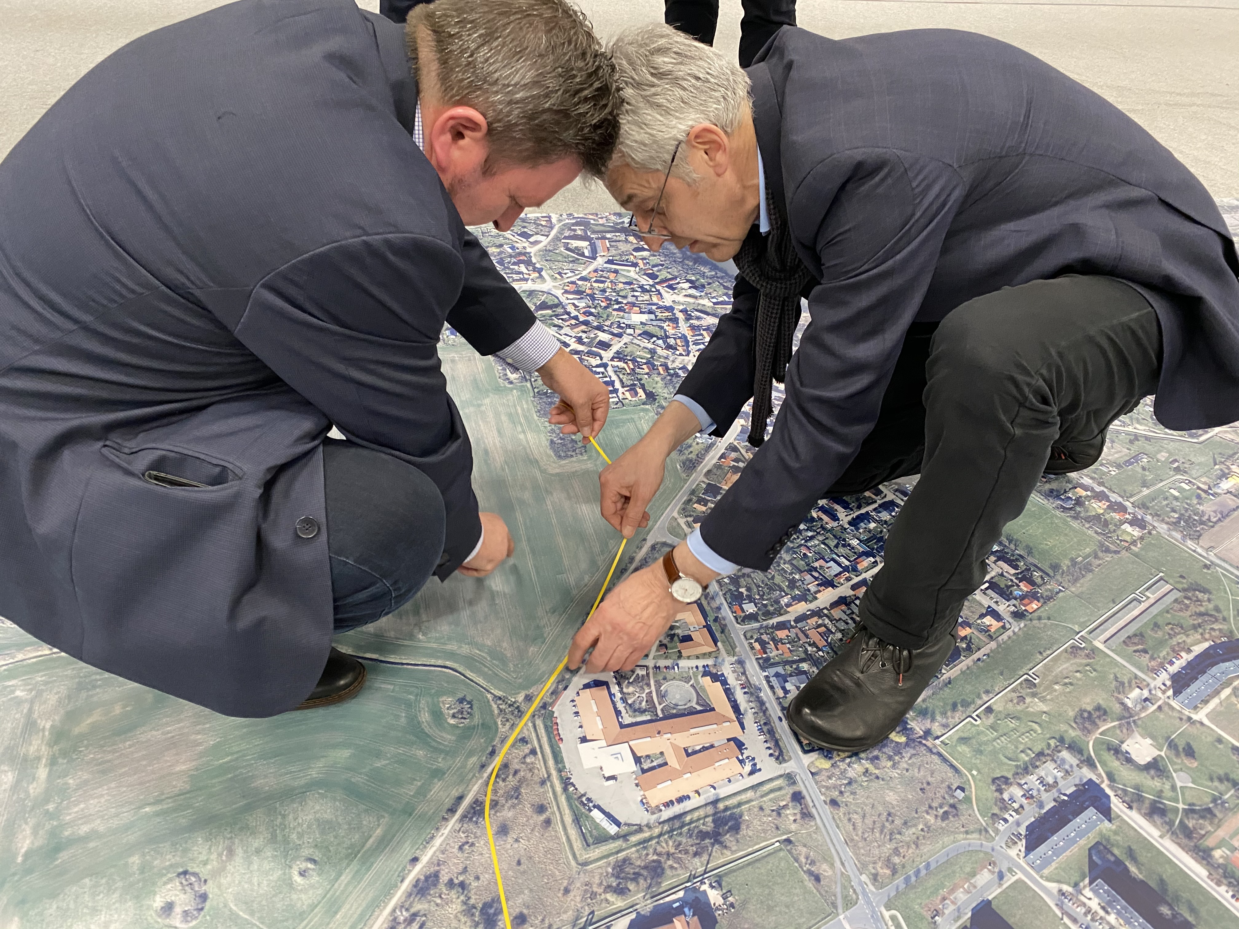 Auf dem Bild sind zwei Männer zu sehen, die auf einem Teppich hocken, welcher Heide-Nord abbildet. Mit einem gelben Klebeband markieren sie auf der Karte, wo die Straßenbahn zukünftig verlaufen könnte.