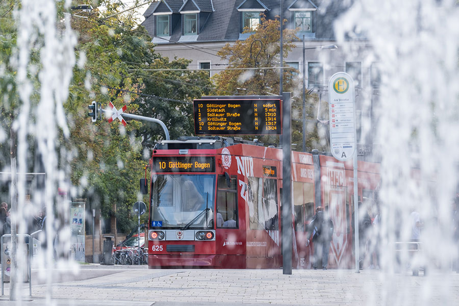 Die rote Straßenbahn der HAVAG fährt direkt auf den Fotografen zu. Die Bahn ist frontal zu sehen und kommt aus einer leichten Rechtskurse. Hinter der Bahn ist ein dunkles Auto. Im Hintergrund sind grüne Bäume mit Laub. Rechts der Bahn ist ein hellblaues Geländer.