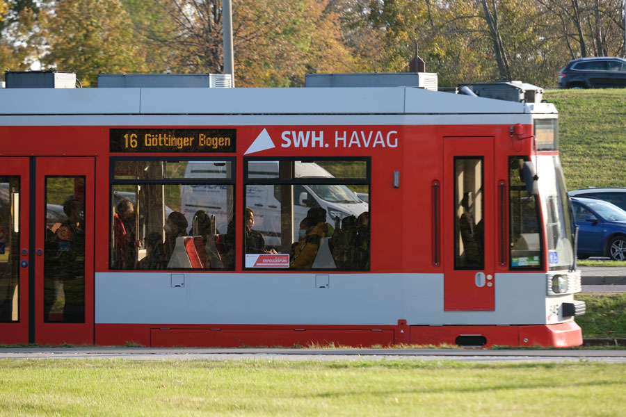 Eine rote Straßenbahn fährt von links nach rechts durch das Bild. Die Straßenbahn ist seitlich zu sehen. In der Bahn sitzen. Vor der Bahn ist ein Grünstreifen zu sehen.