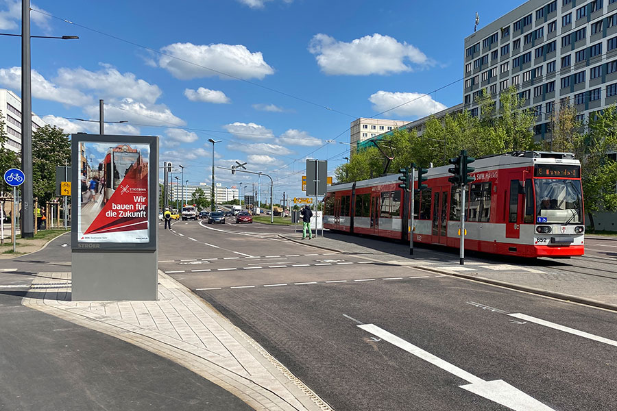 Eine rote Straßenbahn fährt über den Riebeckplatz. Ein Werbeplakat ist zu sehen. Der Himmel ist blau mit kleinen Wölkchen.