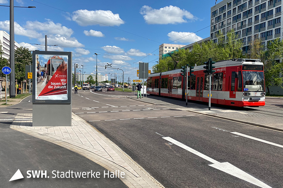 Eine rote Straßenbahn der HAVAG fährt von links nach rechts durch das Bild. Sie überquert den Riebeckplatz. Vor der Straßenbahn ist die Straße mit einem Richtungspfeil. Der Himmel ist blau mit ein paar kleinen Wolken.