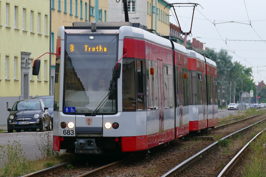 Eine rote Straßenbahn fährt auf Gleisen fast frontal auf den Fotografen zu. Im Hintergrund sind Häuser und parkende Autos.