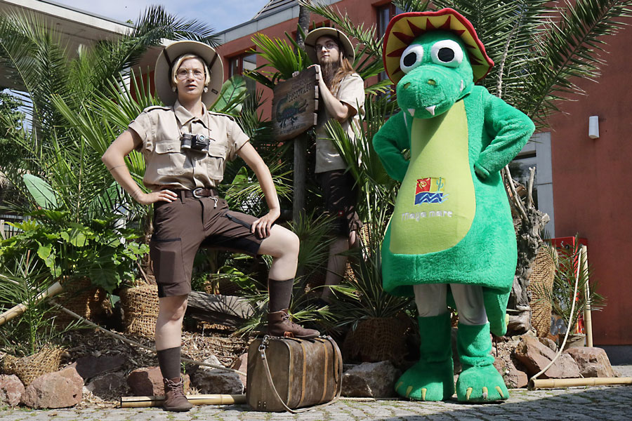Zwei Forscher stehen in voller Ausrüstung neben Mex, dem Krokodil-Maskottchen des Maya mare. Es sind eine Frau und ein Herr. Der Hintergrund sieht aus, als würden Sie in einem Dschungel stehen.