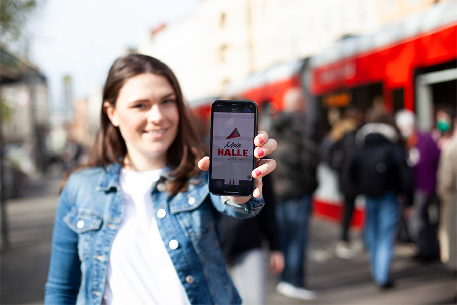 Eine Frau steht auf einem Bürgersteig in der Stadt und hält ein Smartphone in die Kamera, das die App "Mein HALLE Unterwegs" anzeigt. Im Hintergrund sind eine rote Straßenbahn und mehrere verschwommene Personen zu sehen. Die Frau trägt eine Jeansjacke und lächelt.