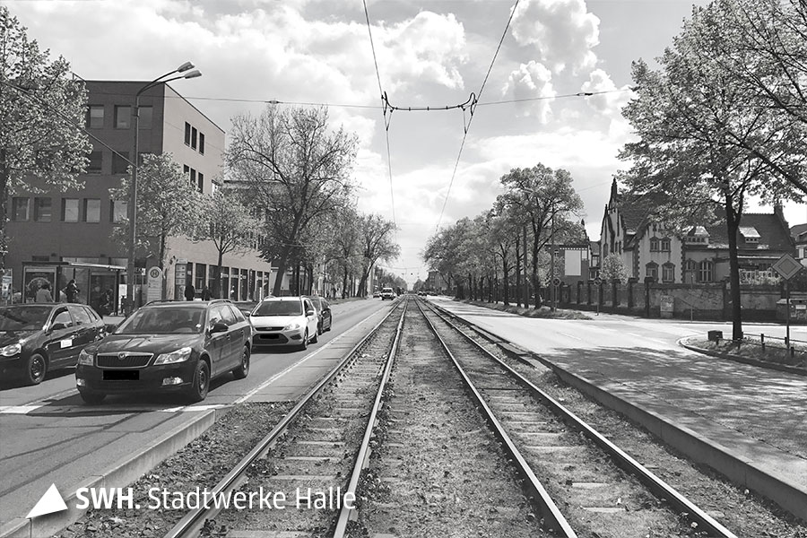 Das schwarz-weiße Foto wurde in der Mitte der Straße zwischen zwei Straßenbahngleisen aufgenommen und zeigt eine lange Straße, auf dessen linker Fahrseite Autos fahren.