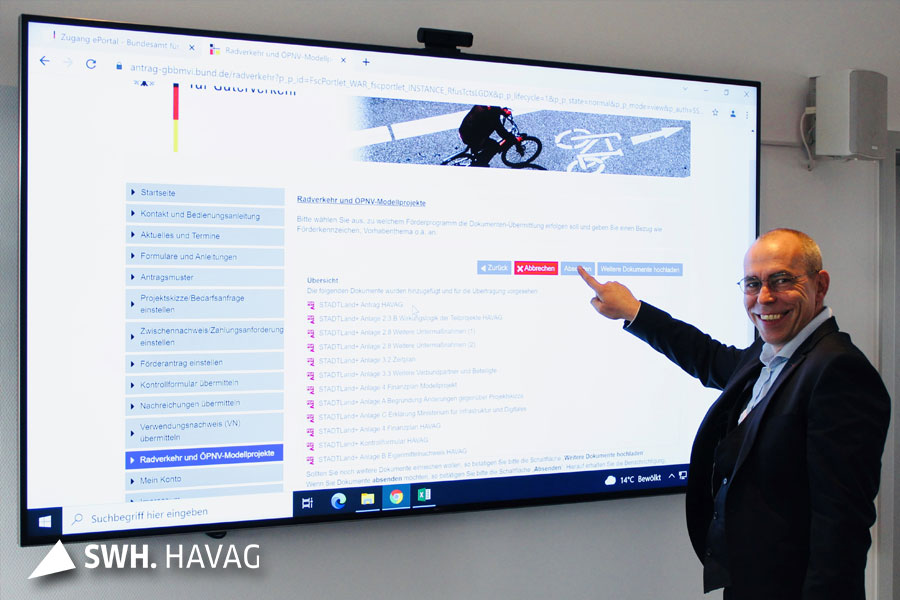 Andreas Völker steht mit einem dunkelblauen Anzug vor einem großen Bildschirm und zeigt mit der rechten Hand auf ihn. Der Bildschirm zeigt die Internetseite der HAVAG.
