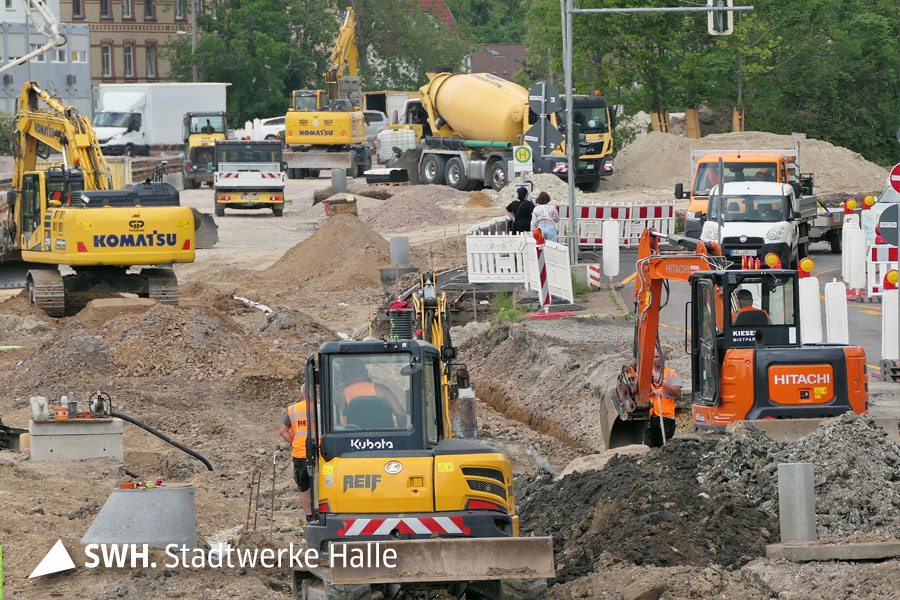 Vier Bagger in gelb und orange, 5 Autos, zwei Transporter und ein LKW auf der Baustelle. Am rechten Bildrand ist eine Behelfsstraße mit Ampel. Dort stehen auch Passanten an einer Haltestelle.