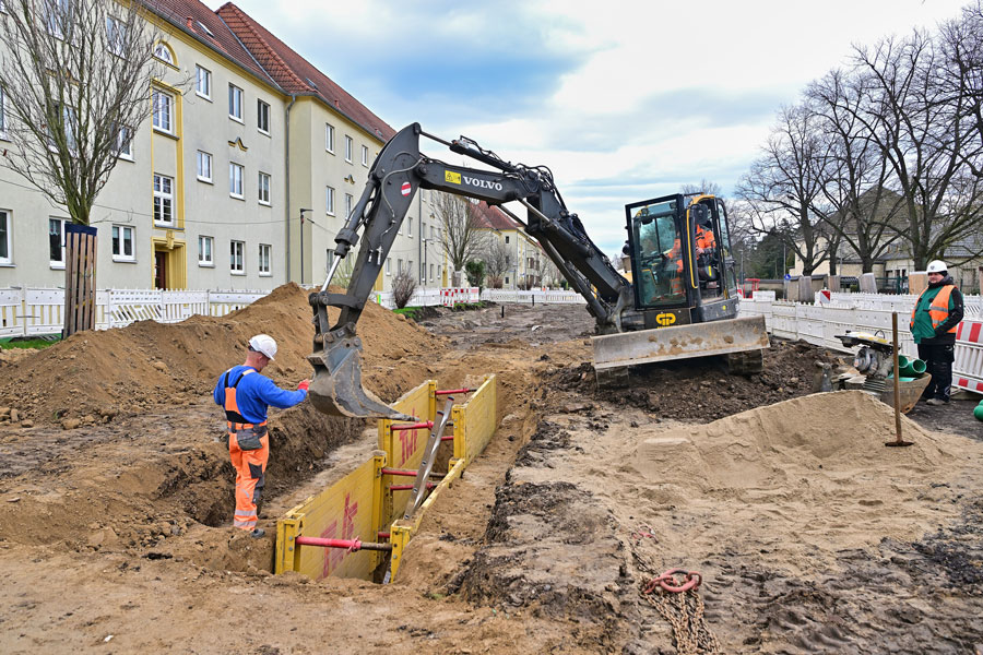 Zwei Männer auf der Baustelle. Ein Bagger baggert. Das Loch wird abgefangen. Im Hintergrund stehen Häuser.