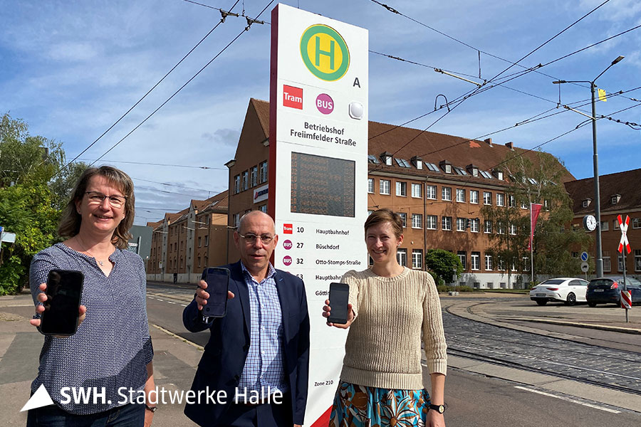 Drei Personen strecken mit ausgestreckter Hand ihr Handy in die Kamera und stehen vor dem Betriebshof Freiimfelder Straße in Halle an der Saale.