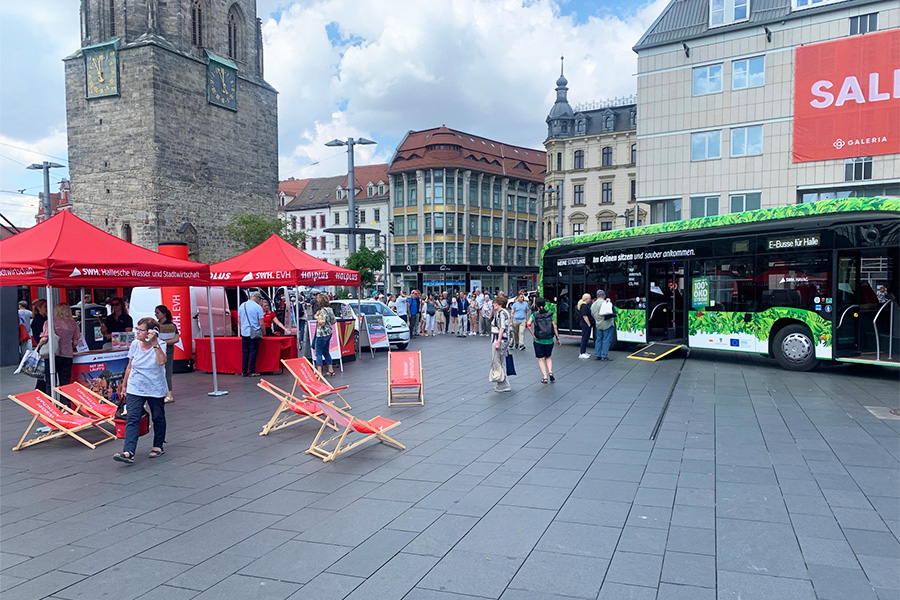 Das Bild zeigt den Marktplatz in Halle, auf welchem mehrere rote Pavillons der Stadtwerke Halle GmbH aufgebaut sind. Unter diesen stehen Informationsstände, an denen Menschen stehen. Daneben sind rote Liegestühle aufgestellt. Im Hintergrund ist ein großer, grüner Elektrobus zu sehen.