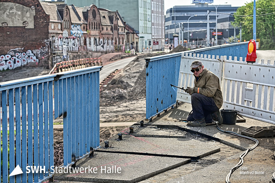 Ein Mann mit Brille und Mütze hockt auf der Brücke. Das blaue Geländer vor ihm fehlt. Er hält ein Arbeitsgerät in der Hand und blickt in die Kamera.