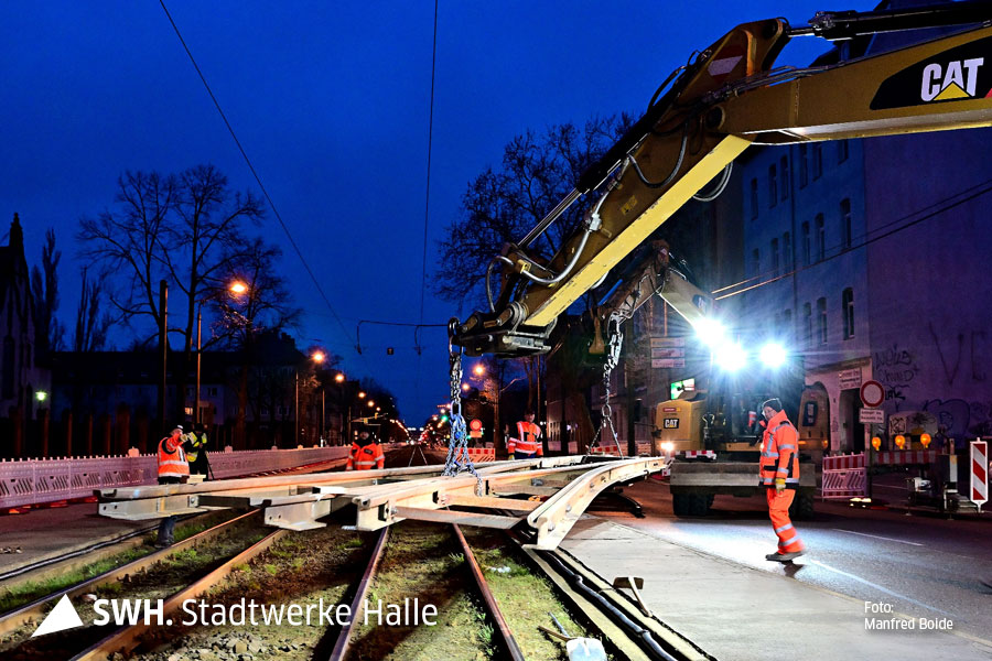 Eine Nachaufnahme: Arbeiter in orangefarbener Arbeitsbekleidung stehen an einem schwebenden Straßenbahngleis. Das Gleis hängt an einem Krank. Der Kranarm ist gelb. Im Hintergrund spenden zwei große Baustrahler Licht. 