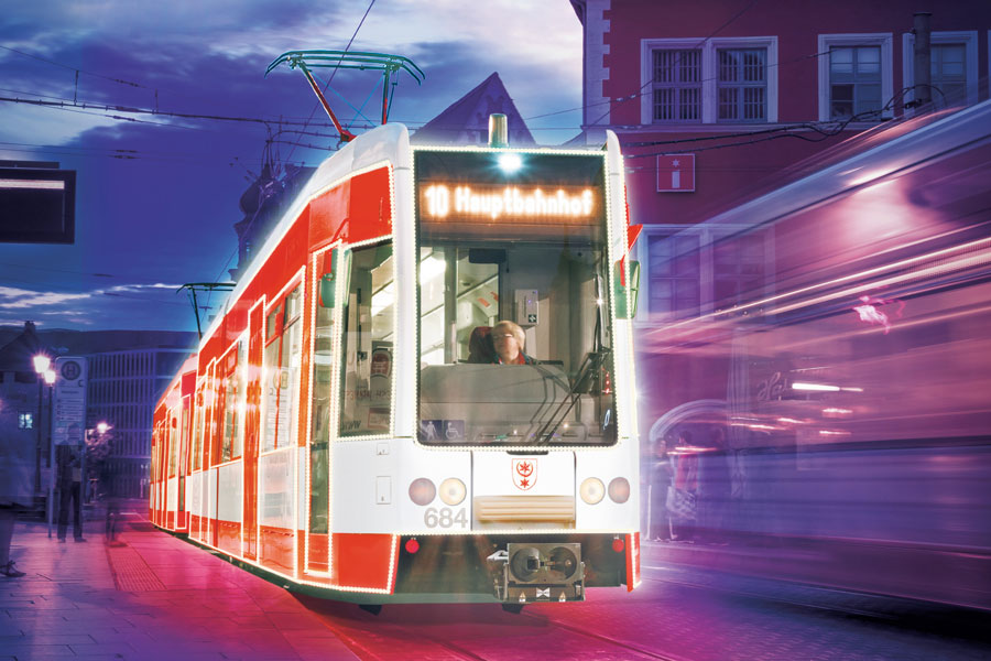 Die Konturen einer roten Straßenbahn leuchten. Es ist später Nachmittag. Die Bahn steht an einer Haltestelle.