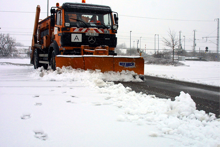 Ein orangenes Winterdienstfahrzeug der HWS auf eine beschneiten Straße, welches Schnee schippt. 