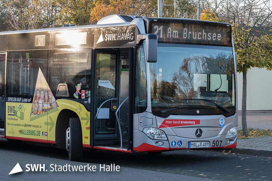 Ein silberfarbener Bus steht an einer Haltestelle. Die Tür ist geöffnet. Die Aufschrift lautet "21 Am Bruchsee".