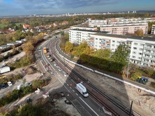 Eine Aufnahme von oben einer Kurve im Böllberger Weg. Ein oranges und ein weißes Baustellenfahrzeug ist zu erkennen. Diese stehen neben den Schienen. Neben den Schienen ist auf der einen Seite eine Straße und auf der anderen Seite noch Baufeld. Im Hintergrund sind Häuserblöcke und Bäume zu sehen.