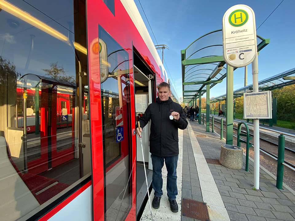 Ein sehbehinderter Mann steht an einer Haltestelle an einer Straßenbahn und zeigt den Handsender.