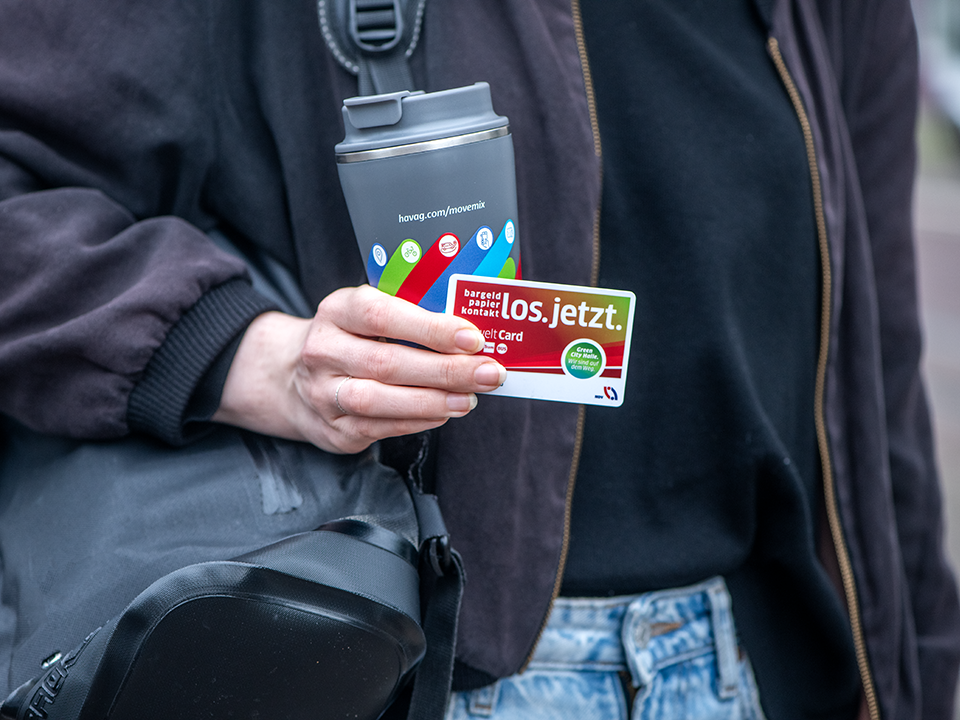 Eine Frau ist mit ihrer Tasche auf dem Weg zur Arbeit und hält einen Kaffeebecher und ihre Chipkarte in der Hand.